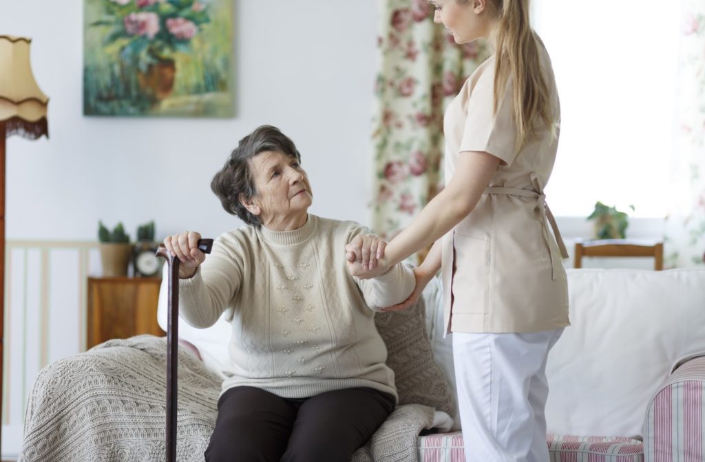 A senior woman standing up off of the couch with help from a supporting nurse