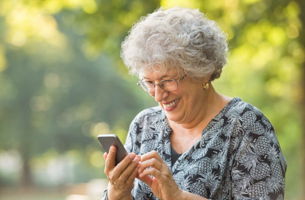 A senior woman standing outdoors smiling while looking at her smartphone.