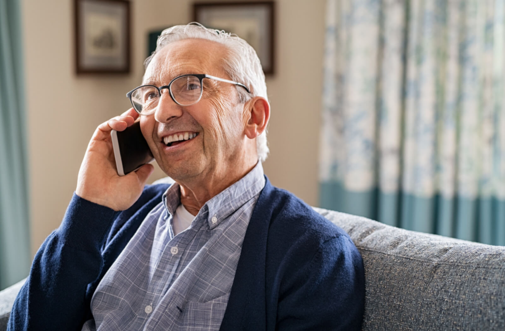 A senior man sitting on a couch with glasses smiling while talking on a cellphone.