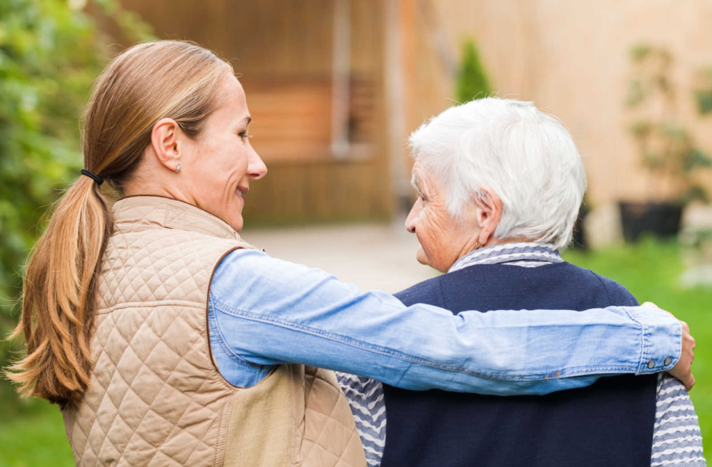 A caregiver walking with a senior woman with dementia while wrapping her arm around the woman's shoulder to comfort her.