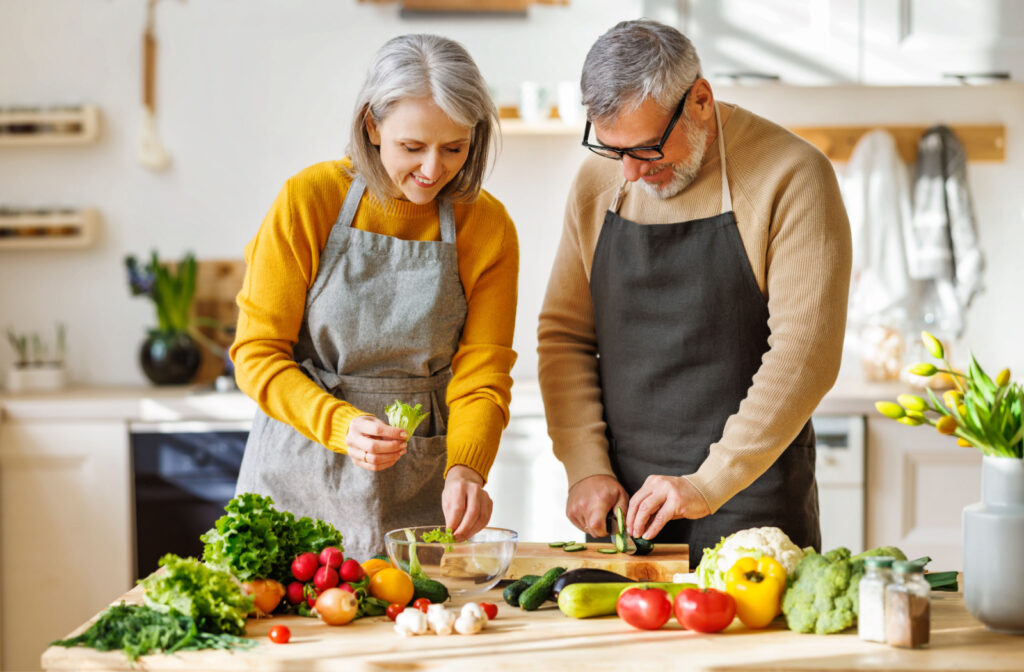 A senior couple wearing aprons and preparing a salad on a kitchen island filled with colorful vegetables