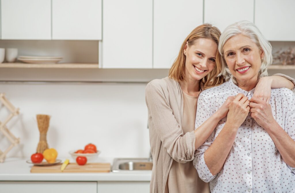 An older adult woman with her daughter hugging her in the kitchen and looking directly at the camera
