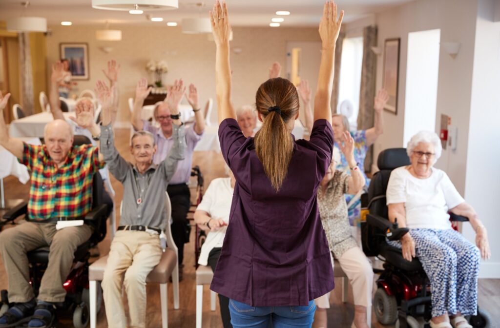 A nurse stands with her back to the camera, leading a group of seniors sitting in front of her through a series of seated stretches