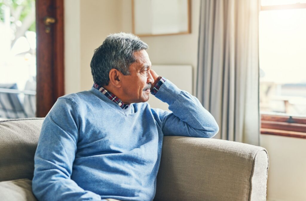 An older adult sits on their couch and looks thoughtfully out the window of their living room