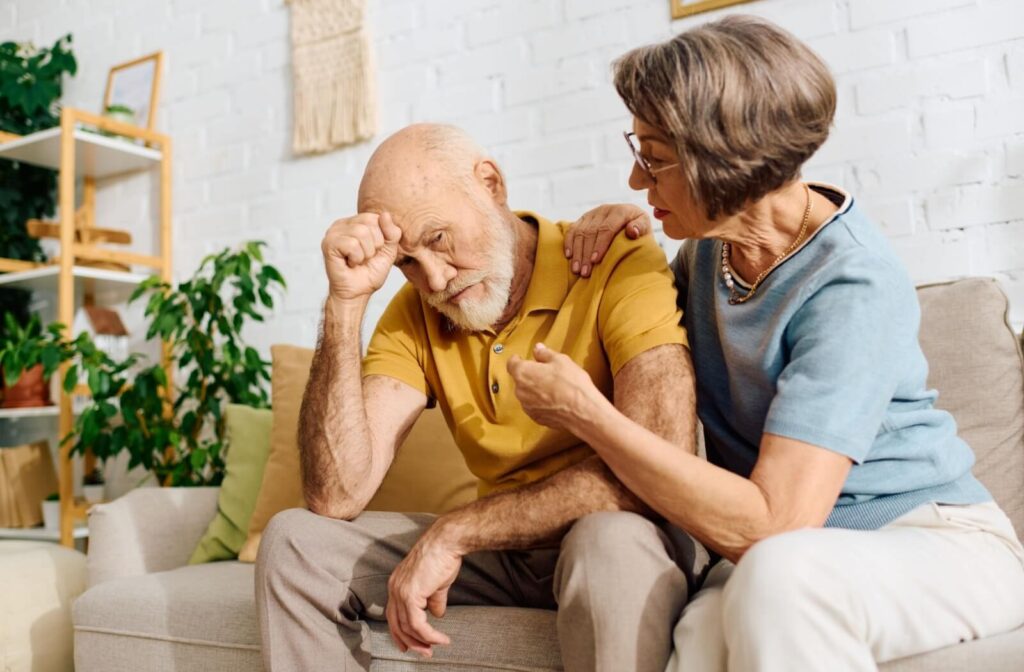 A pair of older adults sits on a couch, one comforting the other as they express distress.